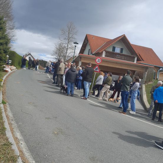 Menschen stehen mit Hunden in einer Reihe auf der Straße, nahe einem Haus mit rotem Dach und 30 km/h Geschwindigkeitsschild.