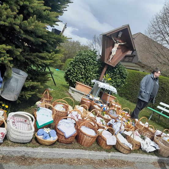 Ein Mann geht an einer kleinen Kapelle mit einem Kruzifix vorbei. Zahlreiche mit Blumen und Tüchern gefüllte Weidenkörbe stehen am Straßenrand.