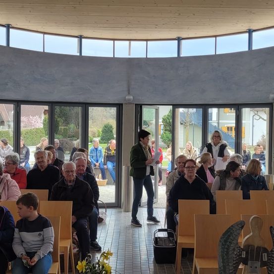 Eine Gruppe von Menschen versammelt sich in einem runden Raum mit bogenförmigen Fenstern. Eine Frau steht in der Mitte, möglicherweise hält sie eine Präsentation. Stühle und ein Tisch sind in der Nähe aufgestellt, und Menschen sitzen und stehen. Draußen gibt es Häuser und Bäume.