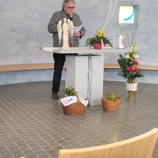 Ein Mann steht an einem Podium mit einem Mikrofon vor einer Wand mit Blumen und einer Kerze. Er hält ein Papier und eine Engelsstatue.
