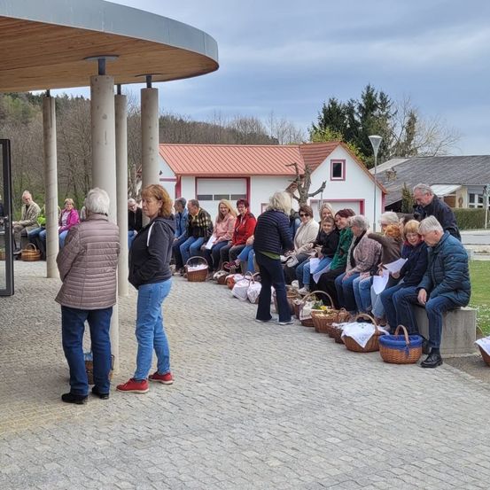 Eine Gruppe älterer Menschen ist vor einem Haus versammelt, einige sitzen auf Betonbänken. Sie halten Körbe. Bäume und ein weiteres Haus sind im Hintergrund.