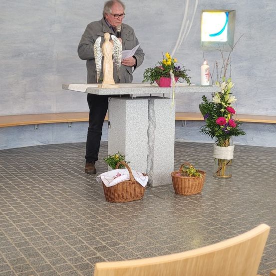 Ein älterer Mann mit Brille steht an einem Podest mit einem hölzernen Engel, hält ein Papier in der Hand und spricht in ein Mikrofon. Es gibt zwei Körbe und Blumen auf dem Boden.