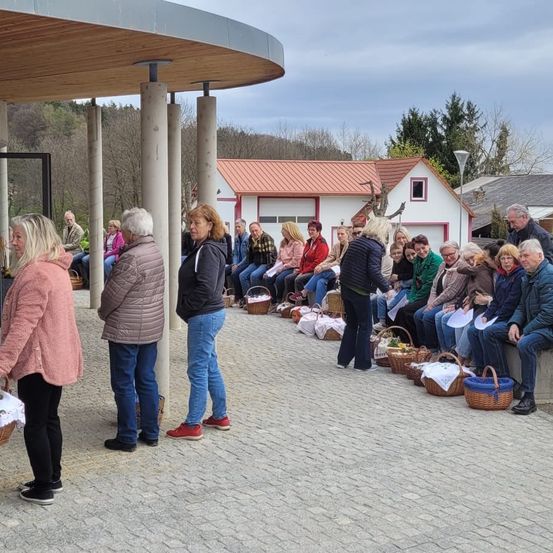 Eine Gruppe von Menschen sitzt auf einer Bank mit Körben vor sich, während andere unter einem Pavillon stehen. Ein Haus mit rotem Dach und Bäumen im Hintergrund.