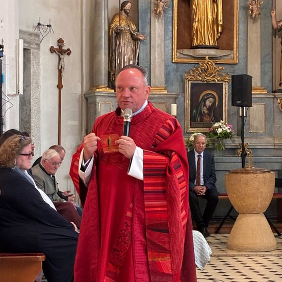 Ein Priester hält eine Rede in einer Kirche, in einem roten Gewand. Menschen sitzen auf Kirchenbänken. Eine Statue und ein Gemälde schmücken die Wand.