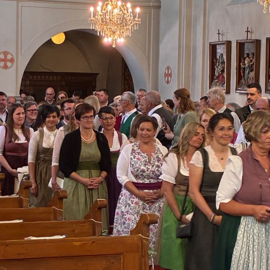 In einer Kirche stehen viele Menschen, wahrscheinlich bei einer Veranstaltung. Die Frauen tragen traditionelle Kleider und Schmuck. Der Raum ist von einer Kronleuchter beleuchtet und hat Holzbänke. Gerahmte Bilder schmücken die Wände.