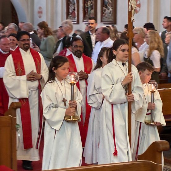 Eine Gruppe von Menschen in weißen Gewändern und roten Schärpen steht in einer Kirche, möglicherweise während einer religiösen Zeremonie. Sie halten brennende Kerzen und stehen vor einem hölzernen Altar.