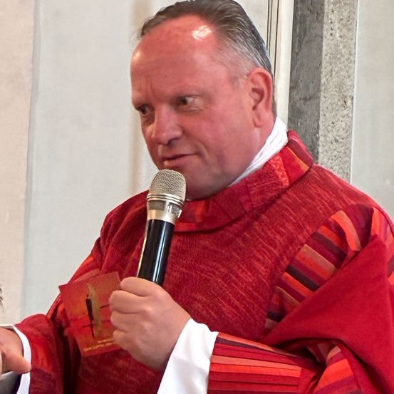 Ein Priester mit grauem Haar hält ein Mikrofon in der Hand und trägt einen roten und weißen Umhang. Er scheint bei einer religiösen Veranstaltung zu sprechen.