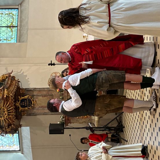 Ein Priester in rotem Gewand segnet zwei Männer in grünen Hemden und Shorts. Sie stehen in einer Kirche mit einem Buntglasfenster hinter ihnen.