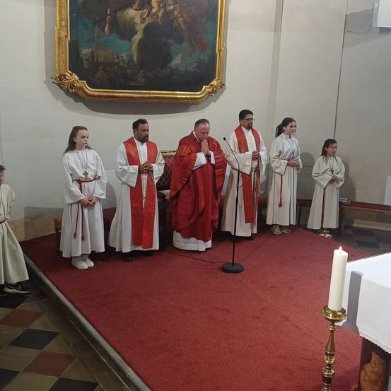 Ein Priester und mehrere Diakone beten vor einem roten Teppich mit einer Kerze auf einem Ständer.