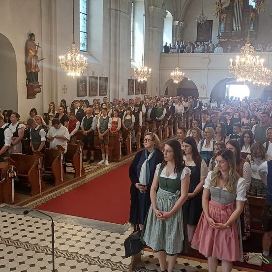 Eine große Menschenmenge in einer Kirche, einige in traditioneller Kleidung, steht aufmerksam. Der Saal ist mit Kronleuchtern geschmückt und weisen kunstvolle Dekorationen auf. Ein roter Teppich führt zur Vorderseite, wo eine Gruppe von Menschen versammelt ist.