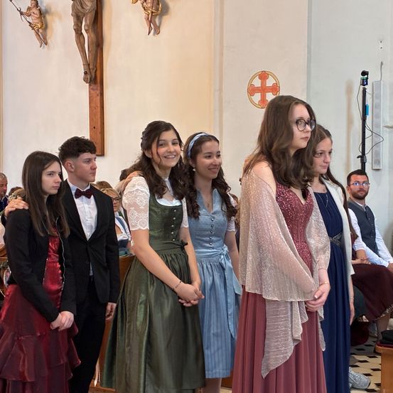 Eine Gruppe von Menschen in formeller Kleidung steht in einer Kirche, möglicherweise während einer Hochzeitszeremonie. Die Frauen tragen Kleider und der Mann einen Anzug. Dahinter sitzen Leute auf Bänken.