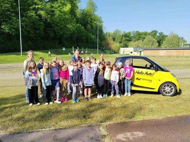Eine Gruppe von Kindern und zwei Erwachsenen steht auf einem Rasenfeld und posiert für ein Foto vor einem gelben Smart-Auto mit dem Text 'Hallo Auto!' auf der Seite.