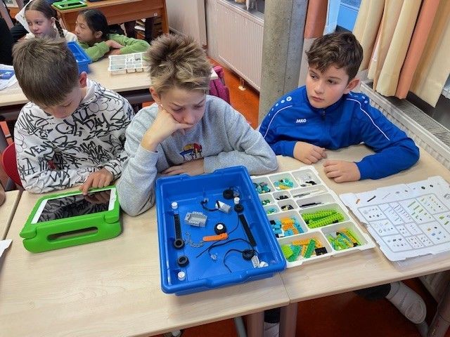 Drei Jungen in einem Klassenzimmer, von denen einer ein Tablet benutzt, während die anderen an einem Projekt mit verschiedenen Lego-Steinen und Werkzeugen auf dem Schreibtisch arbeiten.