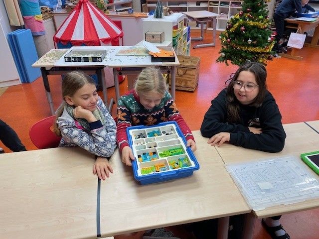 Drei Mädchen sitzen an einem Tisch und beschäftigen sich mit Lego-Steinen, während ein Weihnachtsbaum im Hintergrund zu sehen ist.