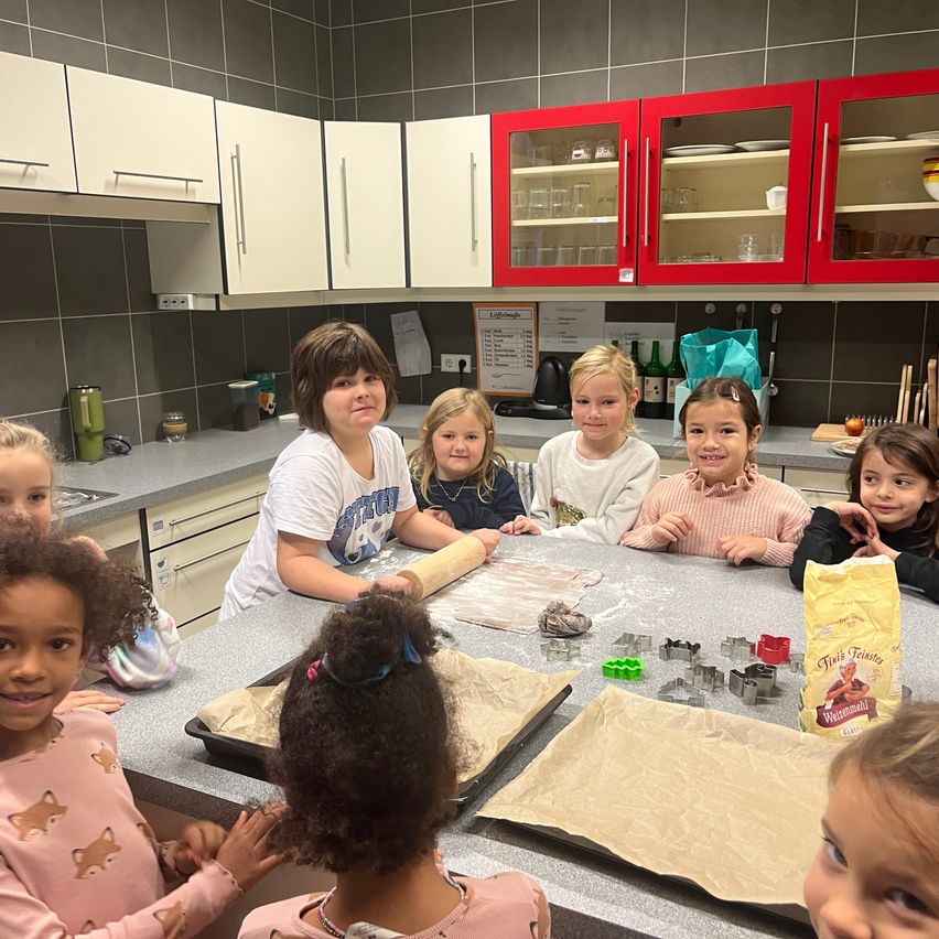 Eine Gruppe junger Mädchen sitzt um einen Küchentisch herum, sie bereiten Teig zu und verwenden Ausstechformen. Ein Mädchen lächelt, während die anderen in die Kamera schauen.