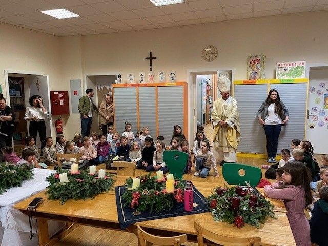 Ein Priester steht vor einer großen Gruppe von Kindern in einem Klassenzimmer, umgeben von Kerzen und Girlanden. Andere Erwachsene sind anwesend, und es gibt Dekorationen und eine Uhr an der Wand.