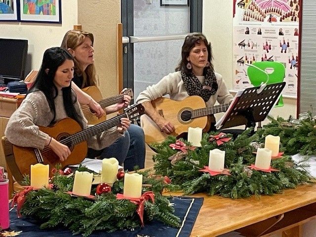 Drei Frauen sitzen auf Stühlen und spielen Gitarren mit Notenständern davor. Der Tisch ist mit einem Weihnachtskranz und brennenden Kerzen geschmückt. Ein Monitor und ein gerahmtes Bild sind im Hintergrund.
