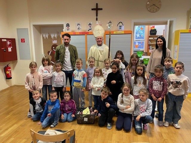 Ein Priester steht mit einer Gruppe von Kindern und zwei Erwachsenen in einem Raum. Eine Box mit Süßigkeiten steht auf dem Boden.