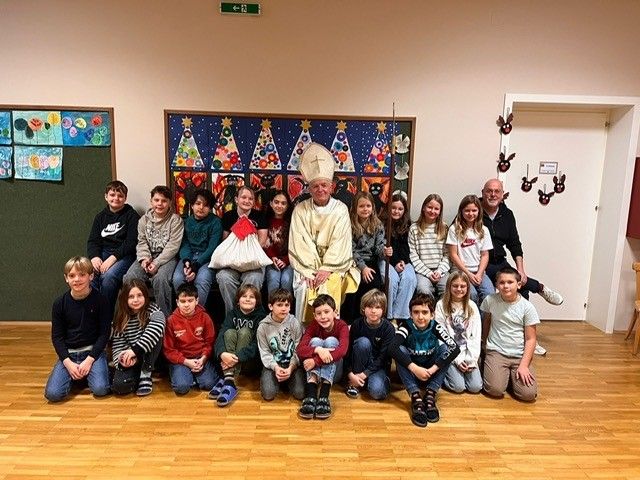 Eine Gruppenaufnahme von Kindern und Erwachsenen, die mit einem Priester in einem Klassenzimmer posieren, mit Weihnachtsdekorationen an der Wand.