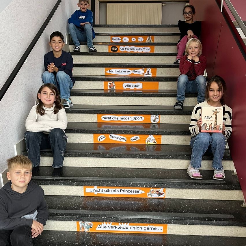 Eine Gruppe von Kindern sitzt auf einer Treppe und posiert für ein Foto. Die Stufen sind mit deutschem Text verziert. Jede Stufe hat eine andere Botschaft. Die Kinder sind lässig gekleidet.