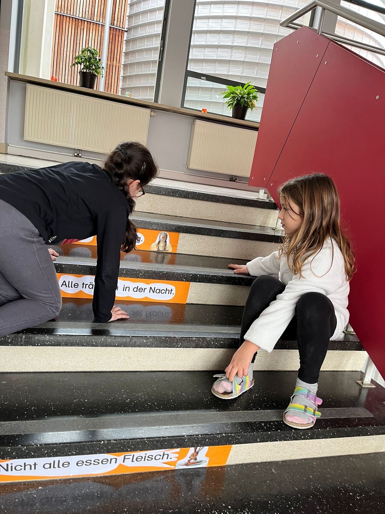 Zwei Mädchen sind auf der Treppe. Eine hilft der anderen mit ihrem Schuh. Auf den Stufen stehen Worte und im Hintergrund befinden sich Topfpflanzen.
