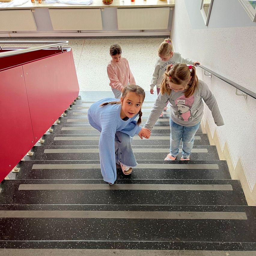 Vier junge Mädchen steigen eine Treppe in einem Flur hinauf. Das Mädchen vorne hält die Hände der hinter ihr stehenden Mädchen. Die Wände sind weiß und der Boden gefliest.