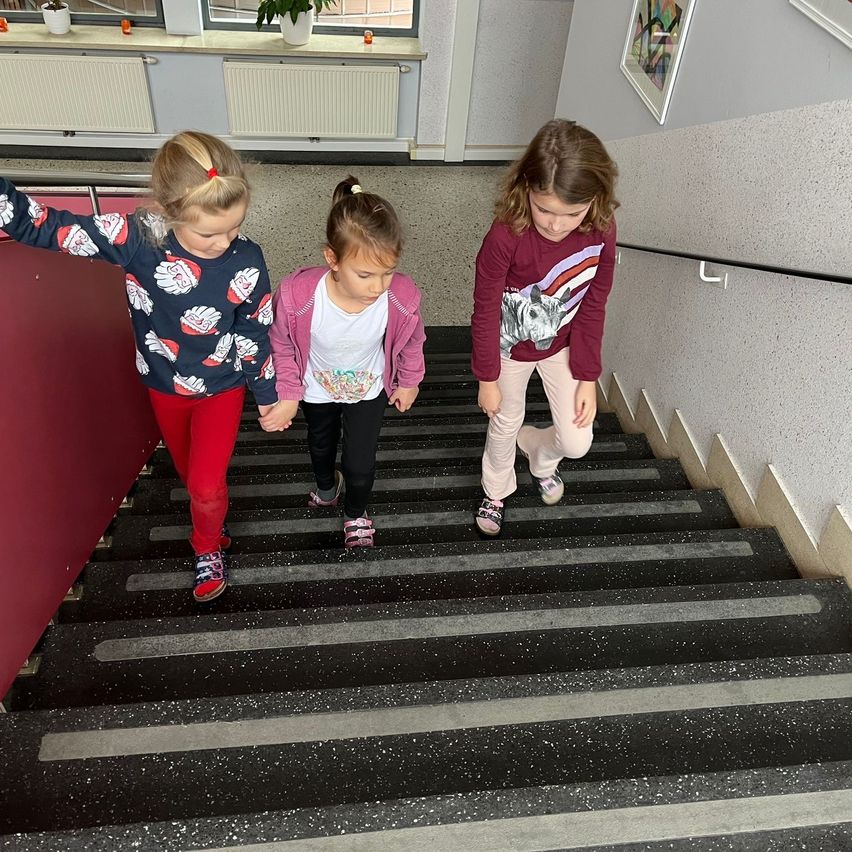 Drei junge Mädchen in unterschiedlich farbiger Kleidung gehen eine Treppe hinauf. Ein Mädchen hält sich an der Handlauf, während ein anderes die Hand des Mädchens neben sich hält.