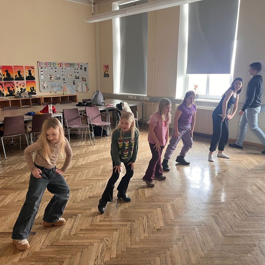 Eine Gruppe junger Mädchen tanzt in einem Klassenzimmer mit Holzböden. Sie tragen Freizeitkleidung. An der Wand befindet sich eine Pinnwand mit verschiedenen Bildern.