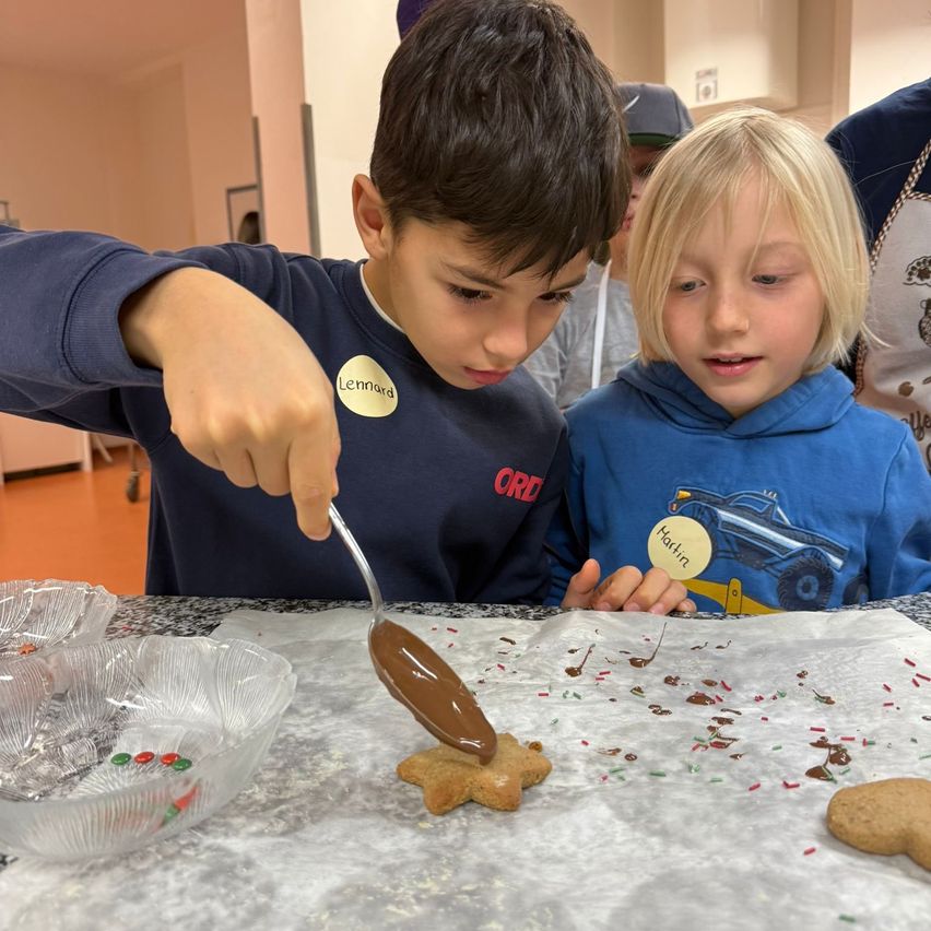 Zwei Kinder, eines mit Namensschild, verzieren einen Lebkuchen mit Schokoladenglasur. Sie sitzen an einem Tisch mit anderen Keksen und Streuseln. Eine Person in einer Schürze steht hinter ihnen.