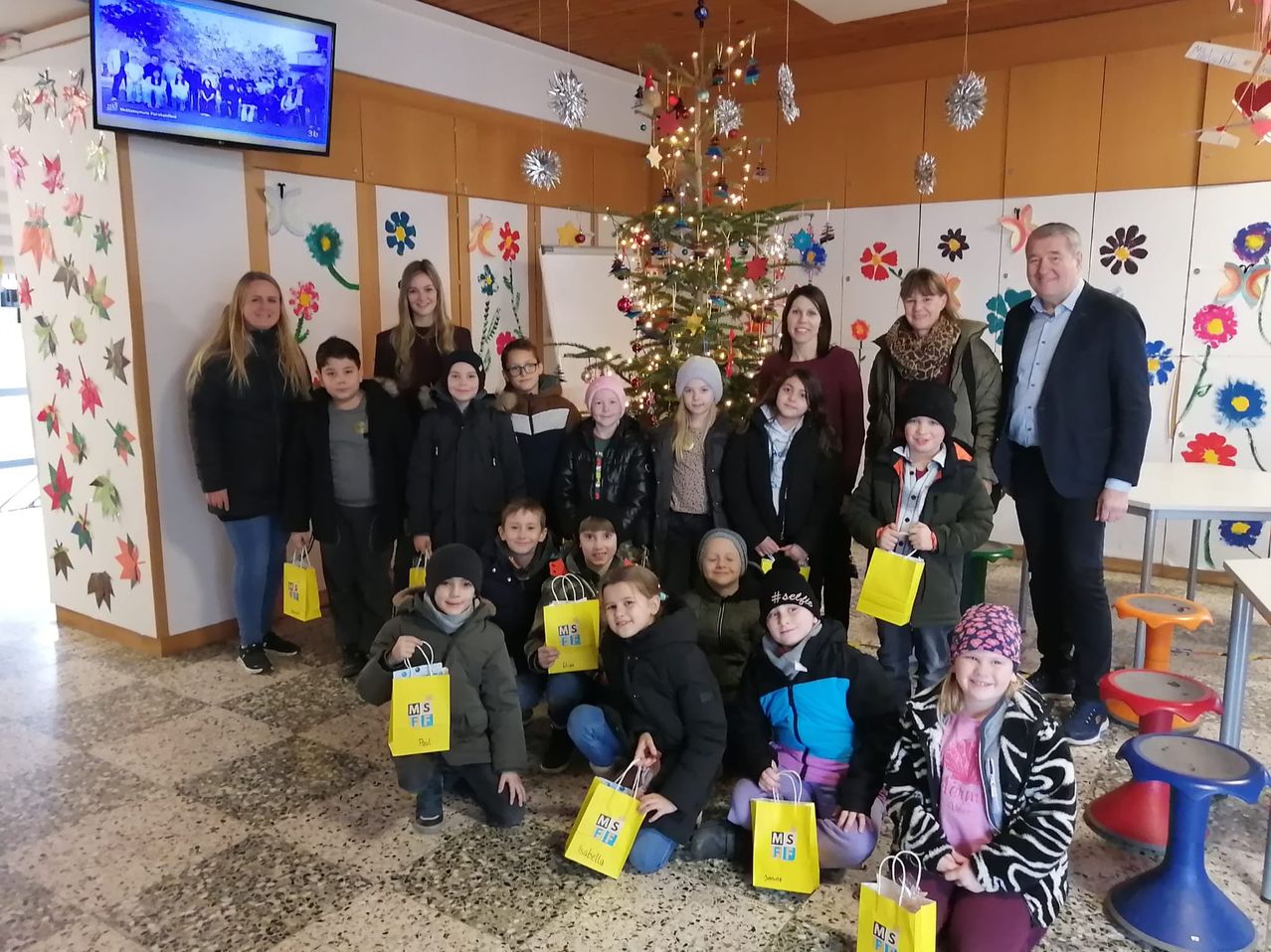 Eine Gruppe von Erwachsenen und Kindern versammelt sich um einen geschmückten Weihnachtsbaum und hält gelbe Geschenktüten. Die Wände sind mit Blumen geschmückt und ein Monitor ist an der Wand montiert.