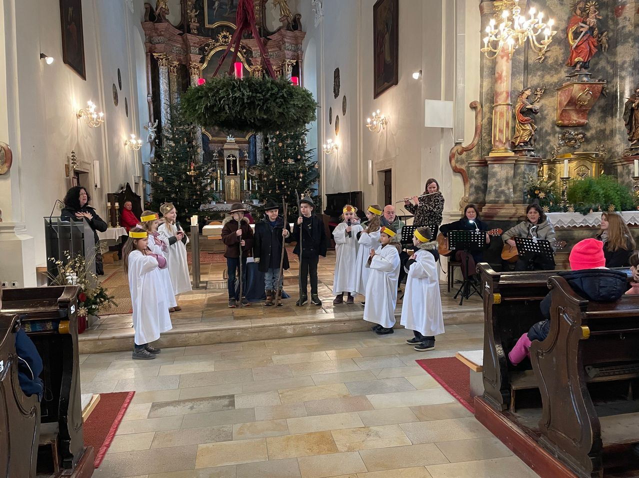 Eine Gruppe von Kindern in weißen Gewändern singt in einer Kirche mit Weihnachtsbäumen und Musikinstrumenten. Eine erwachsene Frau spielt Flöte.