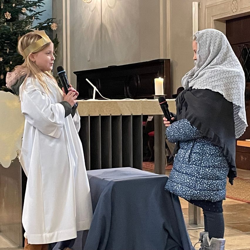 Zwei Mädchen stehen in einer Kirche. Eines hält ein Mikrofon und trägt ein weißes Gewand mit einer goldenen Krone, während das andere ein Mikrofon hält und einen blauen Mantel trägt. Hinter ihnen steht eine Kerze und ein Weihnachtsbaum.