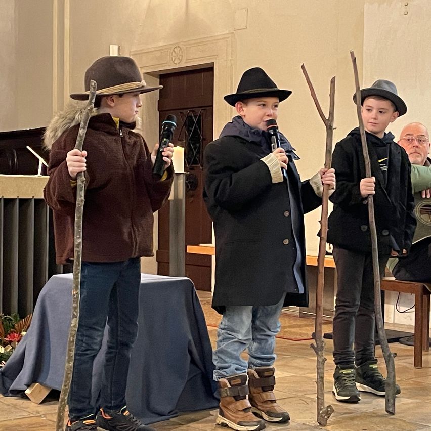 Drei Jungen stehen in einer Kirche, gekleidet in Mäntel und Hüte. Sie halten Stöcke und Mikrofone in den Händen, mit einer Kerze hinter ihnen. Ein älterer Mann sitzt in der Nähe.