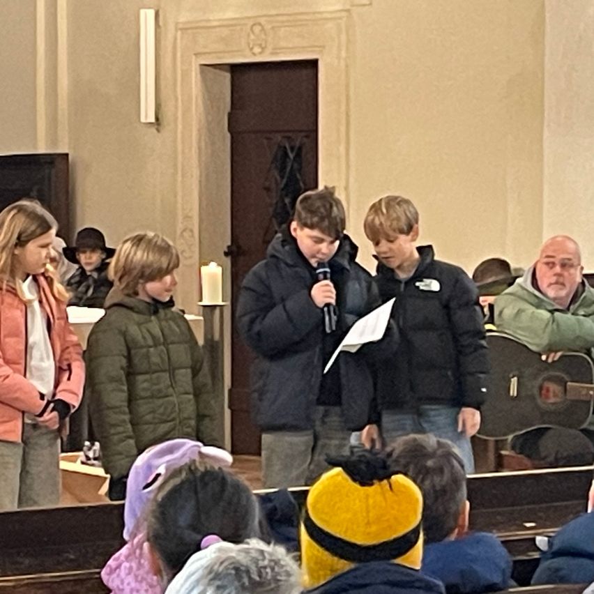 Eine Gruppe von Kindern und Erwachsenen steht in einer Kirche, einer hält ein Mikrofon und liest. Eine Gitarre lehnt an einer Wand hinter ihnen.