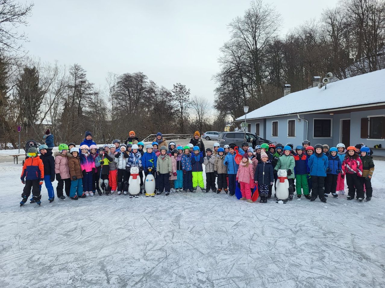 Eine Gruppe von Kindern und Erwachsenen in Winterkleidung posiert für ein Foto auf einer Eisbahn vor einem Gebäude im Hintergrund.