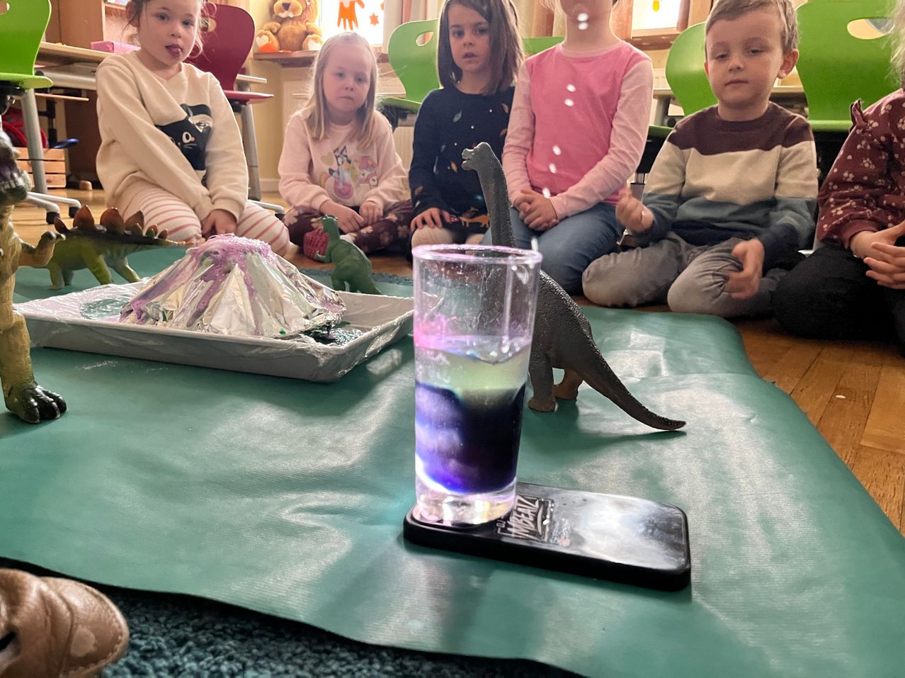 A group of children are sitting on the floor watching a volcano experiment. The volcano is made of foil and has a liquid inside. There are dinosaur toys around the volcano.