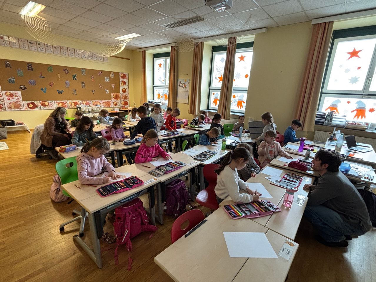 A classroom with children seated at desks, drawing with colored pencils. Adults are seated in the back, observing. The room has large windows, curtains, and a bulletin board with alphabets and drawings.