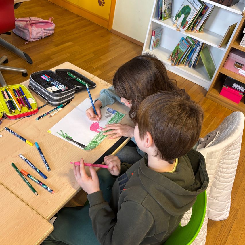 Two children are seated at a desk, engrossed in coloring a drawing. They are surrounded by various colored pencils and markers. A bookshelf with books is visible in the background.