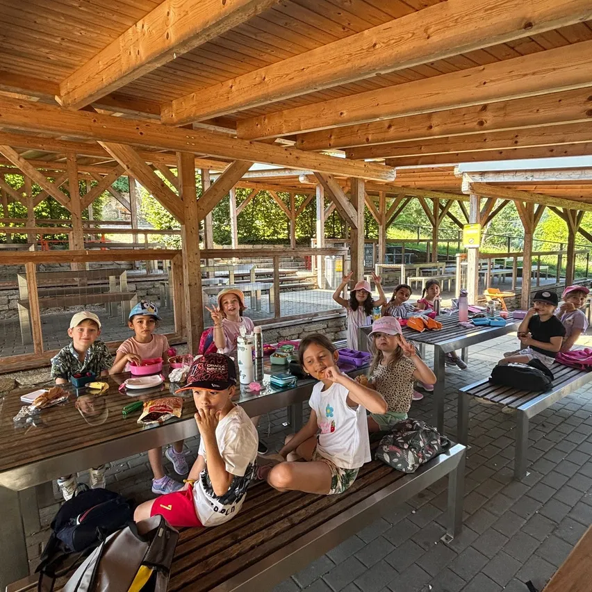 Kinder sitzen in einem schattigen Außenbereich zum Essen an Bänken mit Tabletts, tragen Mützen und Turnschuhe. Einige tragen eine Brille und einer hält einen Becher. Die Holzstruktur hat ein Dach, und Bäume und ein Zaun sind sichtbar.