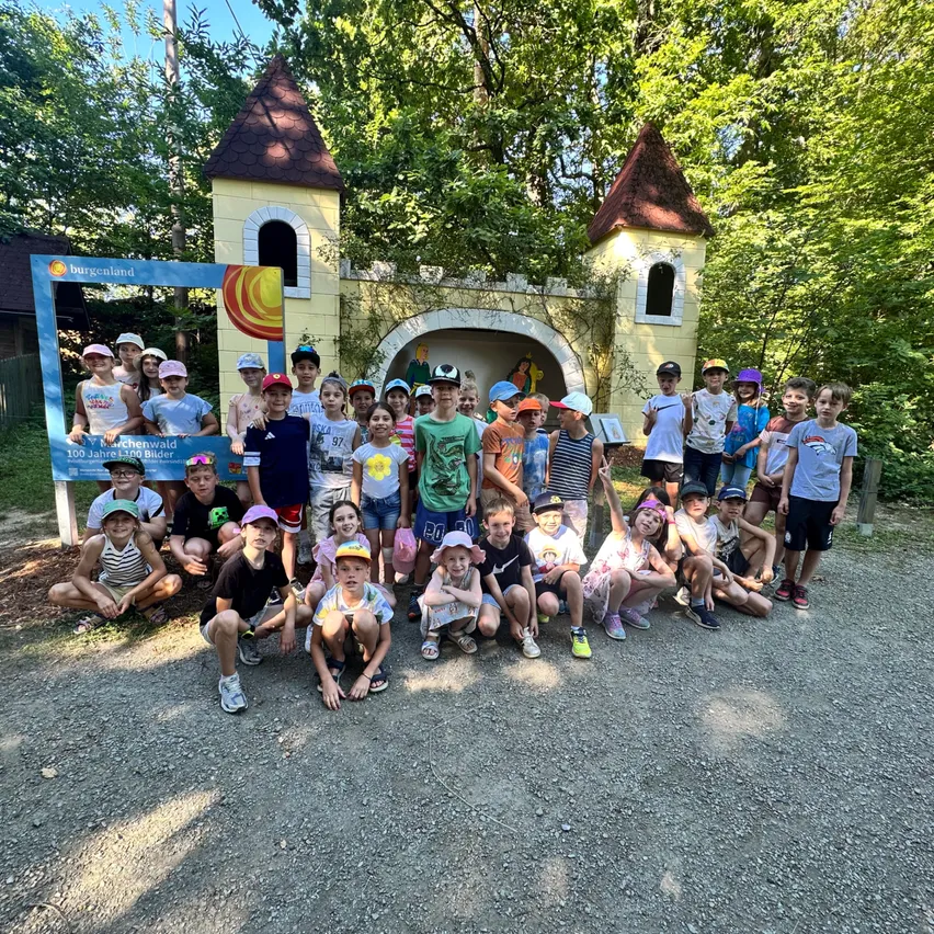 Eine Gruppe von Kindern posiert für ein Foto vor einem burgartigen Spielplatz. Sie tragen Hüte und Turnschuhe. Hinter ihnen steht ein Schild und Bäume.