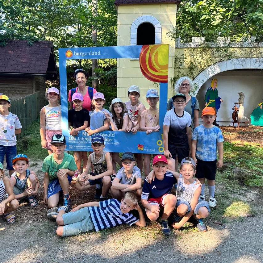Eine Gruppe von Kindern in Hüten posiert für ein Foto vor einem Burgenland-Schild, mit einem gelben Gebäude im Hintergrund.