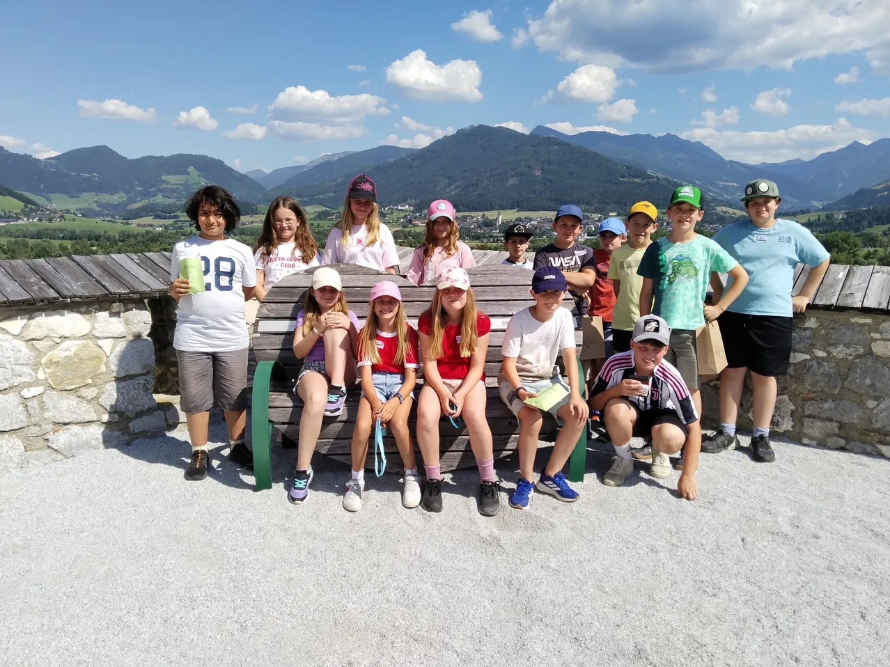 Eine Gruppe von Kindern und Jugendlichen posiert für ein Foto auf einer Holzbank vor einer Bergkette. Die Kinder tragen Mützen und Turnschuhe, und einige halten Tassen. Der Himmel ist blau mit weißen Wolken.