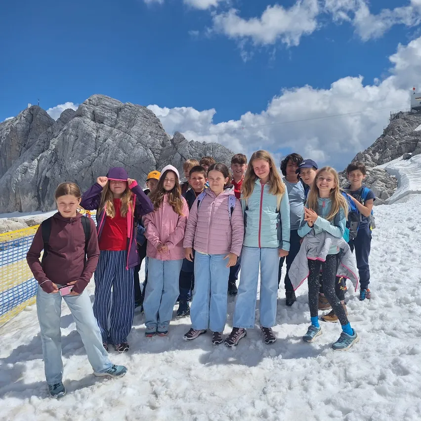 Eine Gruppe von Kindern steht im Schnee und lächelt für ein Foto. Sie sind in Winterkleidung gekleidet, mit Bergen im Hintergrund unter einem teilweise bewölkten Himmel.