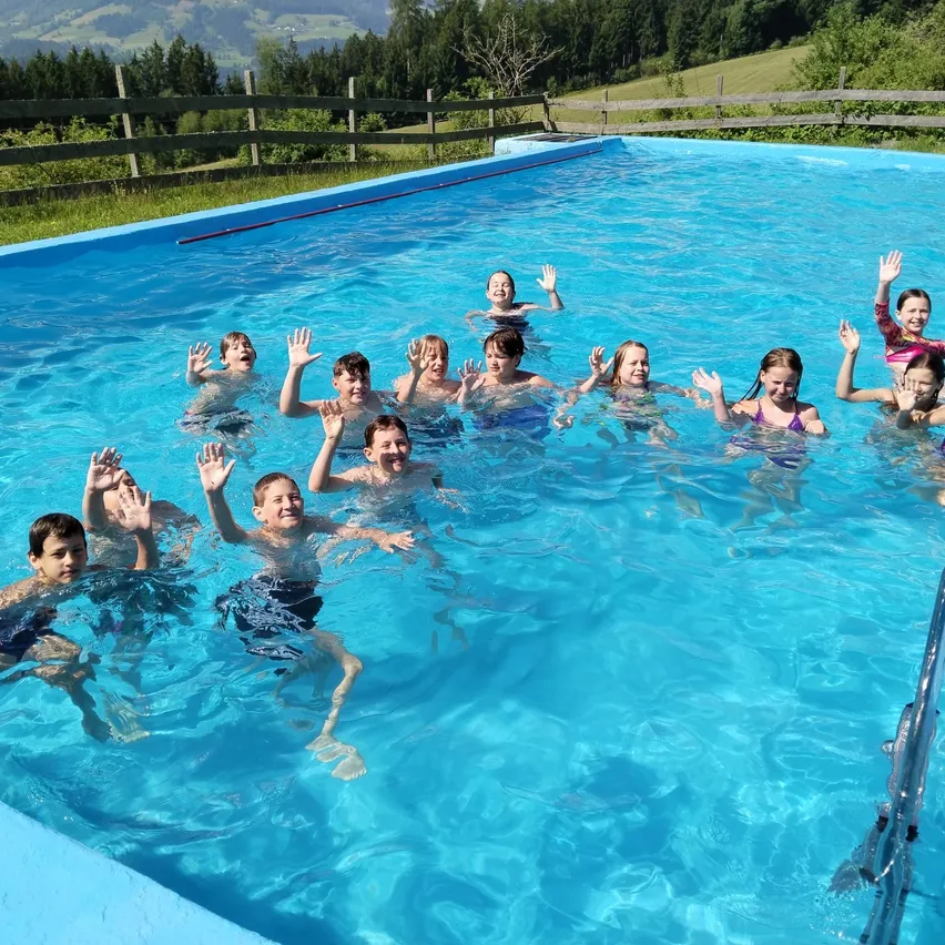 Eine Gruppe von Kindern schwimmt in einem Pool mit klarem blauem Wasser und lächelt. Ein Holzzaun umgibt den Pool, dahinter befindet sich ein Rasen. Bäume und Berge sind im Hintergrund zu sehen.
