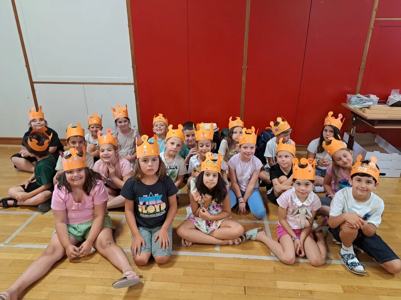 Eine Gruppe von Kindern mit Papierkronen sitzt auf dem Boden in einem Klassenzimmer. Sie sind in Reihen angeordnet und warten scheinbar auf etwas.