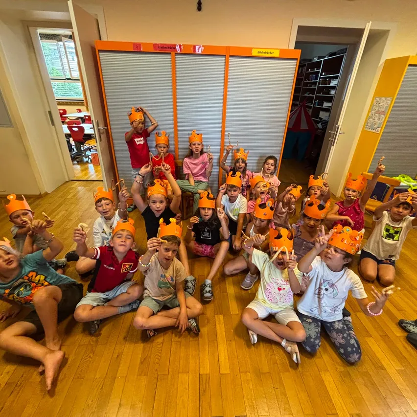 Eine Gruppe von Kindern mit orangefarbenen Kronen auf dem Kopf sitzen auf dem Boden. Sie lächeln alle und posieren für ein Foto in einem Klassenzimmer.