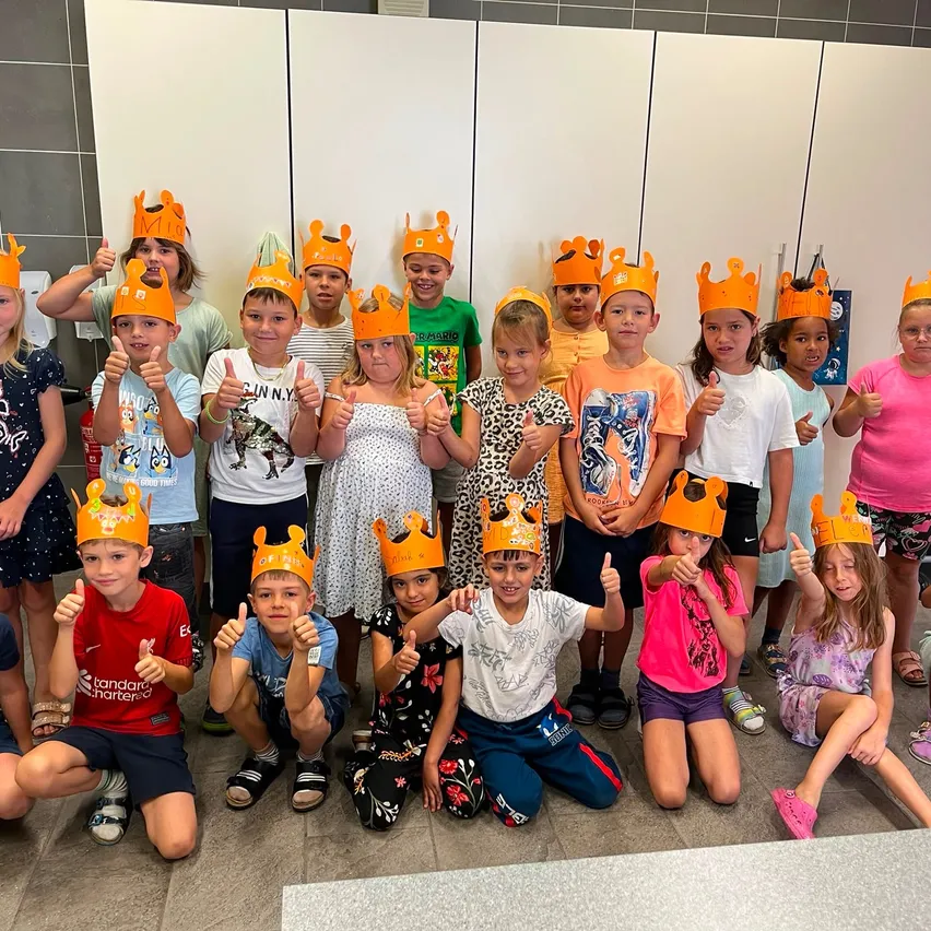 Eine Gruppe von Kindern mit orangefarbenen Kronen posiert für ein Foto. Einige lächeln und geben Daumen hoch. Die Kinder stehen und sitzen auf dem gefliesten Boden. Hinter ihnen ist eine weiße Wand mit gefliester Struktur.