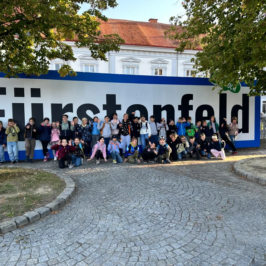 Eine Gruppe von Kindern und Erwachsenen posiert für ein Foto vor einem großen Schild, das 'Fürstenfeld' sagt. Das Schild ist blau und weiß, und im Hintergrund sind Bäume und ein Gebäude zu sehen.