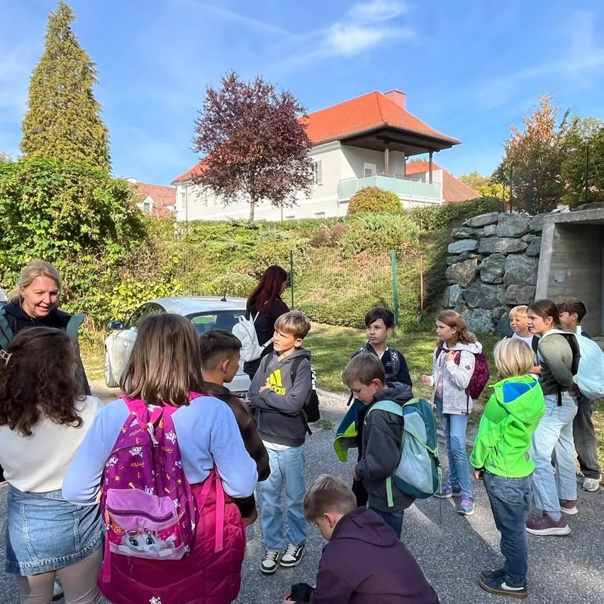 Eine Gruppe von Kindern und einer erwachsenen Frau stehen im Kreis vor einem Haus. Die Kinder tragen Rucksäcke und Turnschuhe.