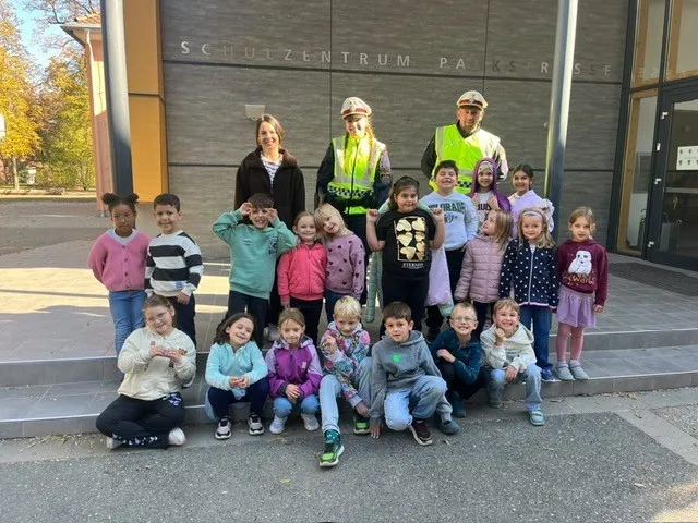 Eine Gruppe von Kindern und zwei Erwachsenen posiert für ein Foto vor einem Gebäude mit den Worten 'SCHULZENTRIUM PAKSRSE'. Die Kinder tragen verschiedene Jacken und Schuhe, und die Erwachsenen tragen reflektierende Westen.